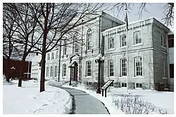 A large grey stone courthouse with arched doorways and tall windows stands amid snow. A cleared walkway curves toward the entrance beneath bare trees.