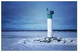 A white cylindrical lighthouse with a green top stands on dark rocks surrounded by snow and frozen water, with a flat winter horizon in the background.