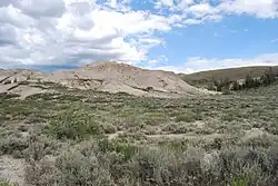 A low gray-tan hill, almost bare of vegetation rising out of the surrounding blue-gray sagebrush.