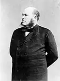 A three-quarters length shot of a Caucasian male with his arms grasping behind his back. He is wearing dark sideburns, a goatie, black bow tie, white shirt and a black coat.