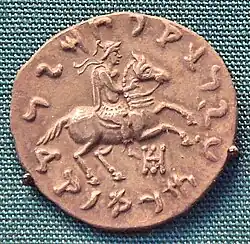 Another coin of Philoxenus. Reverse, showing the king in armour, wearing a crested Boeotian helmet, diadem, chlamys, and boots, while riding a galloping horse. He is also making a blessing gesture with his right hand.