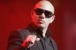 Picture of a man in a suit, wearing sunglasses and holding a microphone in his hands against a red backdrop.