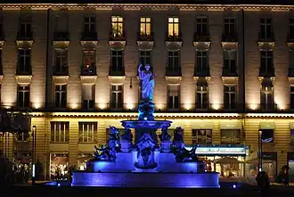 The fountain illuminated in blue, standing out against the beige and ochre façades.