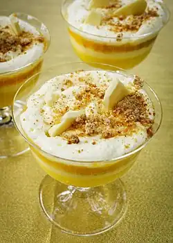 A close up image of a Platinum Jubilee pudding#. It focusses on a white cream top with Amaretti crumb, over a yellow toned layered trifle. Shards of white chocolate are also on top of the cream.