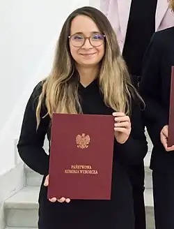 Photograph of Zawisza in her mid-thirties. She is wearing a black dress and round glasses, holding a red book titled "National Electoral Commission". Her hair is long, and both blonde and brunette.