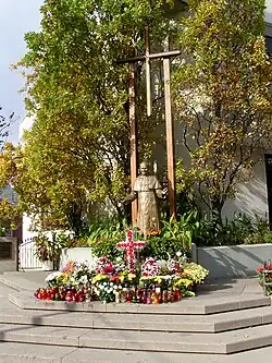 Statue of man in robes standing in front of large cross, with flowers and candles at his feet