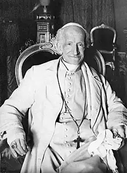 An elderly Pope Leo wearing a white suit, a long cross-necklace, a ring, a white zucchetto, and a large ring, sitting in an ornate chair, holding a cloth, and smiling
