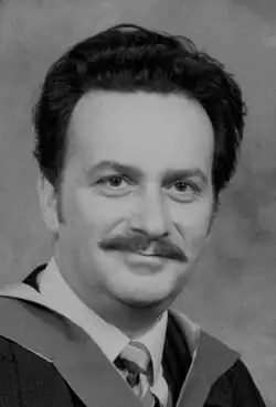Neville Bower circa 1985. Black and white portrait photograph of an Anglo-Indian man with dark hair and a moustache wearing formal academic attire including a masters gown and hood.