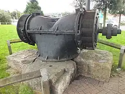 A large iron water turbine displayed supported on a group of stones, with a small unreadable metal plaque, in a retail park with surroundings of lawn, pavement, and low rail fences