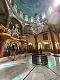 A spacious hall crowned with a painted blue dome surrounded by colorful stained-glass windows. Red marble columns support the arches, giving the space balance and dignity.
