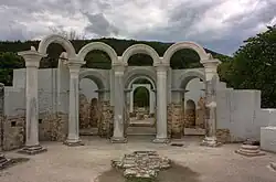 Five marble columns near the walls of a partially preserved building. A gate and a small monument are in the background.