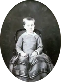 Photograph of a young Isabel with hair gathered at the back and wearing earrings seated on a small, cane back armchair