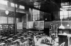 A black and white image of a large room with may chairs in front of a desk and charts. There are bars on the windows.