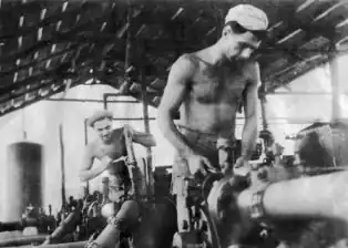 Black and white photograph of two shirtless men fixing equipment in a pumping station