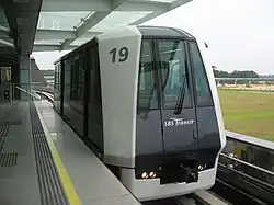 A white and dark blue Mitsubishi Crystal Mover train at the elevated platform of Punggol LRT station. The train, numbered 19, is stationary with its doors closed, and beyond the platform, an open grassy field and some construction or distant structures are visible.