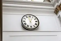 Close-up photograph of a wall-mounted clock with a white face and black Roman numerals, on a pastel-yellow wall.