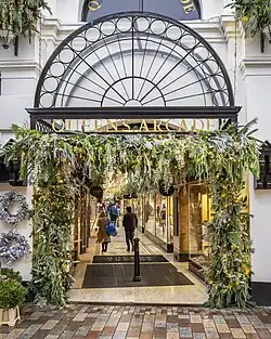 A two-storey building fronting onto a pedestrian street. The building is painted pastel-yellow and features black-framed shop fronts. An ornate black-framed semi-circular canopy protrudes above the entrance to the arcade, with gold lettering spelling out "Queen's Arcade".
