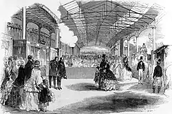 An artist's impression of the inside of a train station crowded with people underneath a long roof. One group of these is obviously identifiable as Queen Victoria, Prince Albert, and one of their chlidren. Two men bow their heads opposite them.