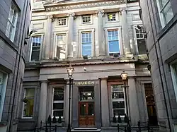 White two-storey building of Classic architecture, with Doric pillars on the ground floor, Ionic pillars on the second, and two Victorian gas lanterns illuminating the entrance; the word "QUEEN" in gilt lettering over the entrance