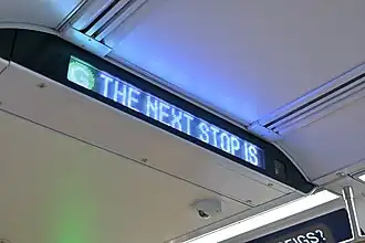 An LED sign on the ceiling of a subway car displaying "The next stop is", and a ceiling-mounted security camera