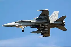 Photo of a modern fighter aircraft viewed from below. The aircraft's landing gear are extended, and bombs and fuel tanks are attached to the lower surface of its wings.