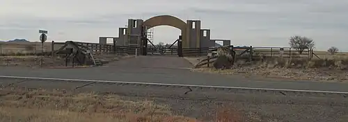 A driveway off of a paved road in a level grassy area; there are two layers of gates and a cattle guard crossing the driveway