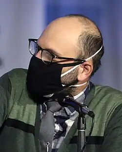 a balding man wearing a black mask stares to his right