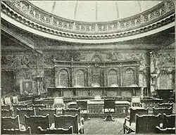 Interior of the courtroom. There are seats facing a judge's bench with elaborately decorated panels behind it. Above is a domed roof.