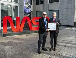 Two men standing in front of a large red NASA "worm" logo sculpture holding a framed certificate