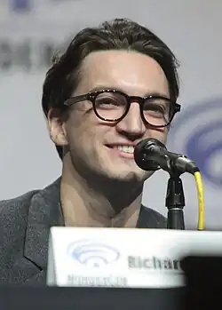 Richard Harmon wearing glasses, looking to the right of the camera and smiling.