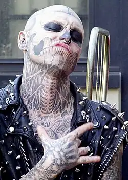 A man covered in skeleton-like tattoos on his face, neck, and hands, wearing a spiky leather vest.