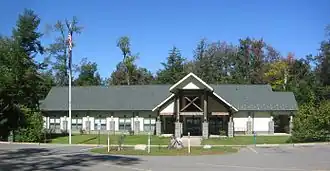 Photo of a modern one-story building with five windows and a two-story entrance with wooden framing and "Ricketts Glen" in metal letters. There are trees on three sides, with a grassy lawn and parking lot in front, and a bright blue sky.