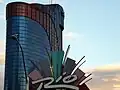Distant ground-level view of an all-glass tower; the tower has several setbacks near its roof. The glass is mostly a dark blue color, but some windows are tinted red to form a stripe down the side of the building. A prominent "Rio" sign is visible in the foreground.