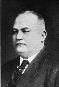 A balding man wearing a black jacket, vest, and tie and white shirt