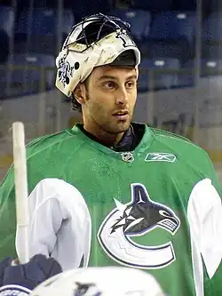 An ice hockey goaltender with his mask pulled off of his face looking forwards. He wears a green jersey with a logo of a stylized orca in the shape of a "C".