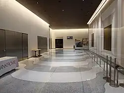 View of the lobby, facing north, along the passageway on the western side of the elevator core. There are white and gray pavers on the floor. To the right is a wall with the artwork "Portals", which includes alternating bands of white and brown glass surrounding a set of bronze and nickel plates.