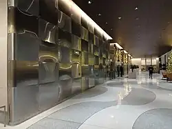 View of the lobby, facing east along the southern side of the elevator core. There are white and gray pavers on the floor. The walls of the core have panels arranged in a checkerboard pattern.