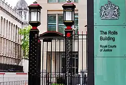 The entrance to the Rolls Building. A pair of black iron gates with large lanterns on top, next to a large glass sign saying, "The Rolls Building".