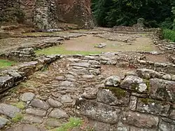 Ruined foundations of buildings, some patches of ground covered in cobblestones; at the far and near ends of the foundations the stonework is built up to around a metre tall; a castle wall can be seen in the background left.