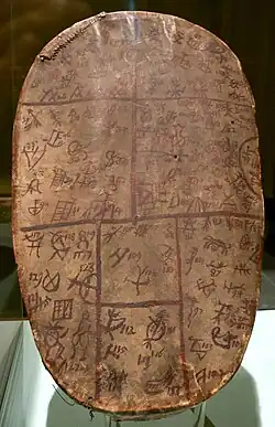 An oval leather drum with faded brown and red symbols covering the surface, divided into sections by painted lines, shown in a museum display.