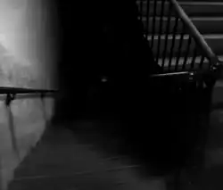 A very dark staircase in which the bottom is not visible with a floating, deformed human face faintly visible in the distance