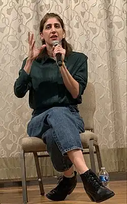 Photo of Abed sitting in a chair and speaking into a microphone. She has pulled back shoulder length brown hair and wears a dark green button up shirt, denim capris, and black ankle boots.