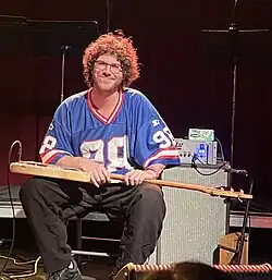 Sam Wilkes in 2024 wearing a jersey and sitting on a chair with an electric bass on his lap, in front of an amp.