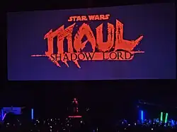 Actor Sam Witwer on stage, covered in red light, with the text "Star Wars: Maul – Shadow Lord" on the screen behind in red
