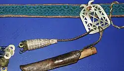 A woven belt with a patterned band, metal buckle, cords, and attached needle cases and small metal disks, shown in a museum display.