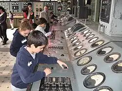 Children fiddling with knobs on a long flat grey metal surface with large dials facing them