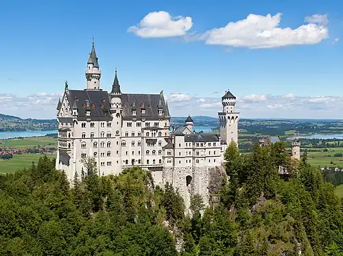 A castle of fairy-tale appearance sitting high on a ridge above a wooded landscape. The walls are of pale stone, the roofs are of steep pitch and there are a number of small towers and turrets.