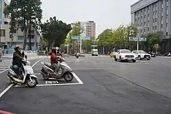 Scooters waiting for the traffic signal within a dedicated hook turn zone marked as a white rectangle, in Kaohsiung