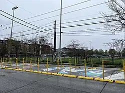 A mural is on the street surrounded by yellow barricades with soccer fields in the background. The mural's M has a white background, covered in splashes of blue, teal, yellow, and purple paint. Part of an E,S and A are shown: the S is colored in red, black, and white.