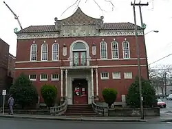 Two-story red brick building with small rectangular first-floor windows, stairs to red doors on the first floor, and tall arched windows on the second floor with a small porch over the first-floor door and a pointed facade above.